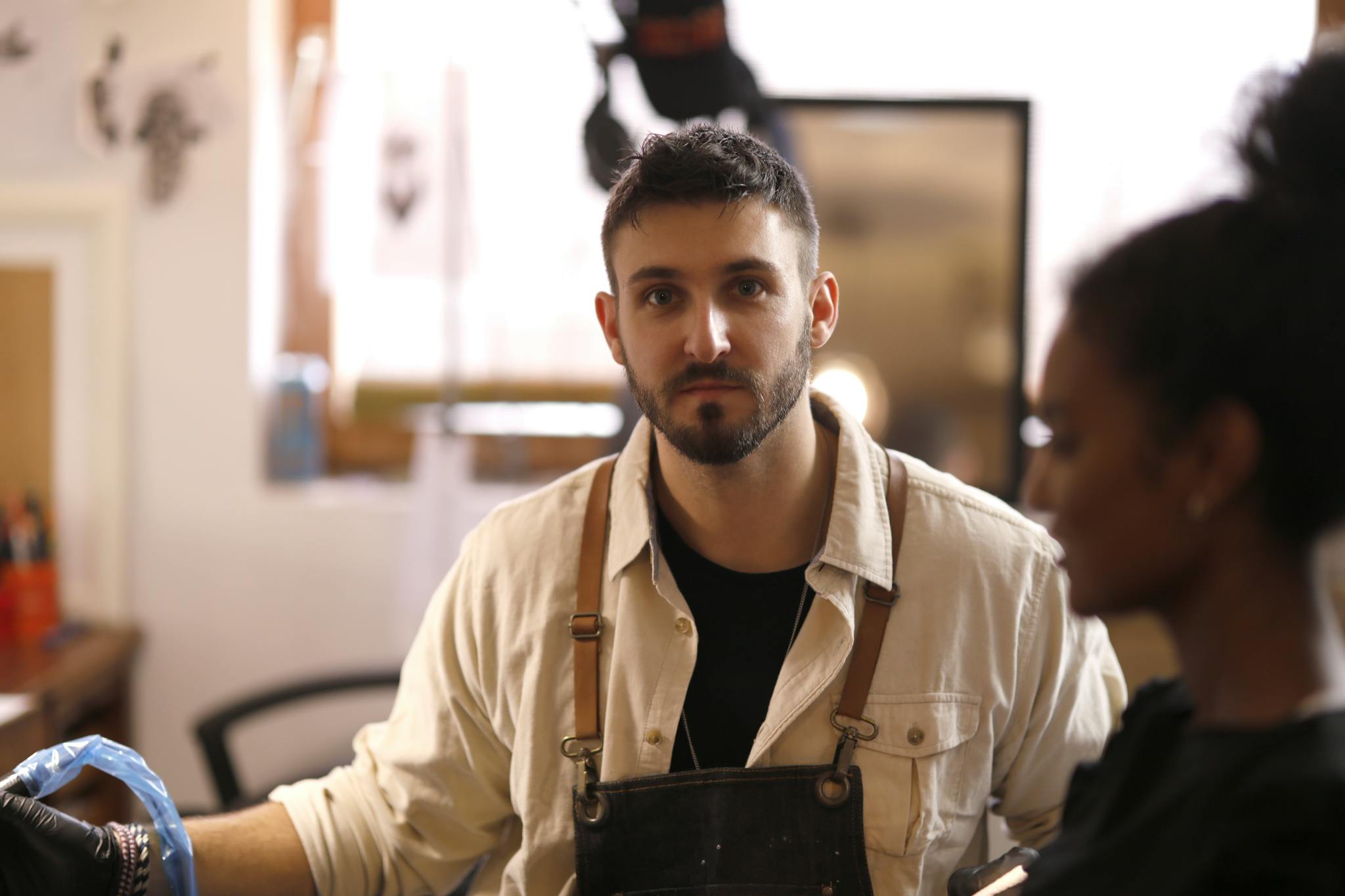 Positive young bearded tattooist wearing apron and black latex gloves working with client in modern salon and looking at camera